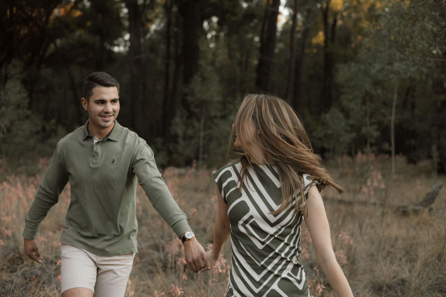 A smiling man in an olive green polo leads a laughing woman in a striped dress by the hand through a sunlit forest.