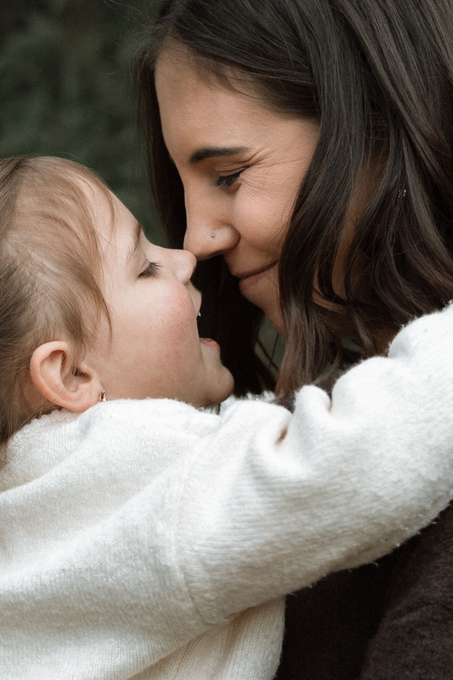Smiling mother in a dark sweater touches noses with her toddler daughter in a white fuzzy jacket during an outdoor family session.