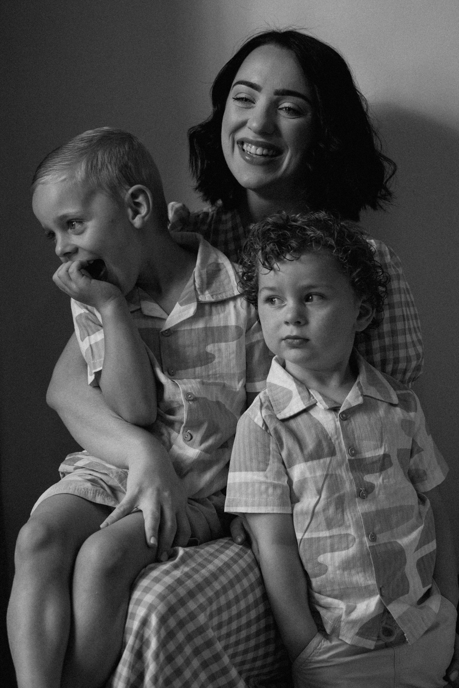Black and white portrait of a smiling woman with two young boys in matching camouflage-print shirts, one covering his mouth with his hand.